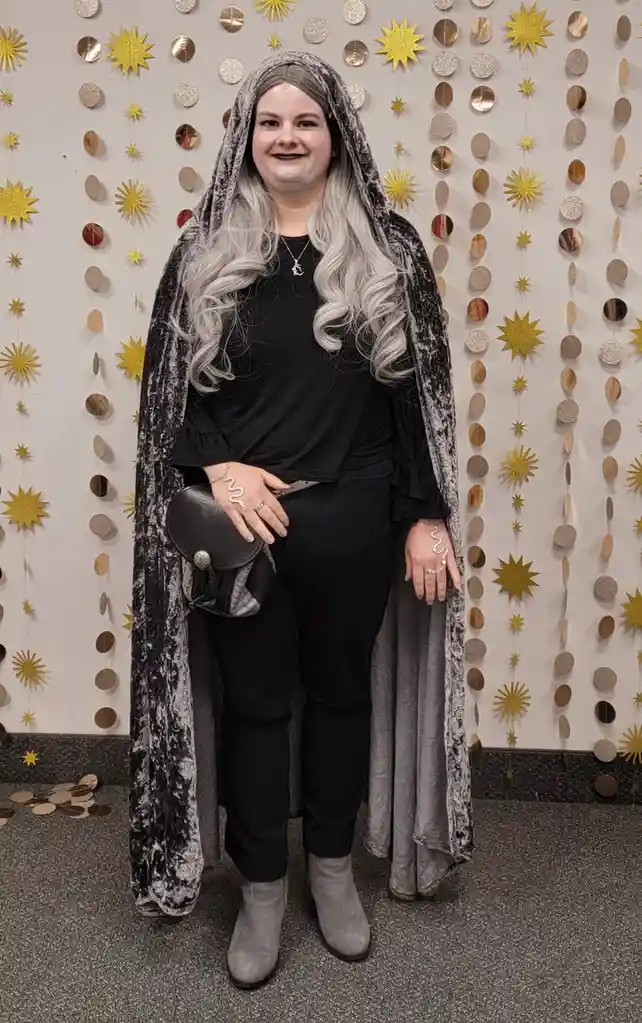 A person with long silver hair and pale face paint stands in a dark velvet cloak against a wall decorated with gold and…