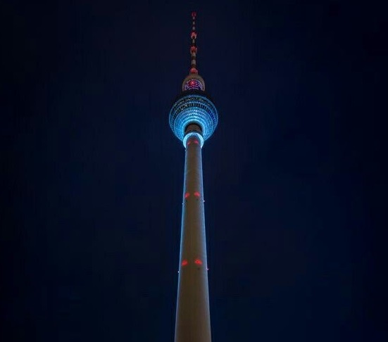 A low-angle nighttime photo of the Berlin TV Tower, its main shaft and spherical observation deck illuminated with blue and…