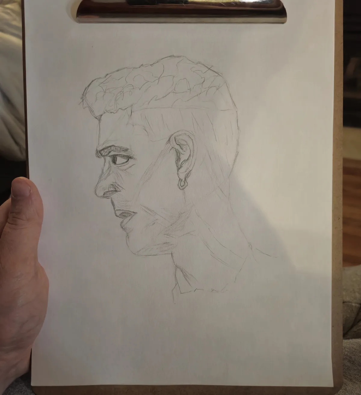 A hand holds a clipboard displaying a rough pencil sketch of a person's head and shoulders in profile. The drawing shows a…