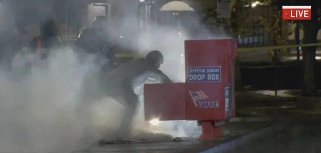A red official ballot drop box is engulfed in smoke and flames at night, with a person in dark clothing and a hooded head…