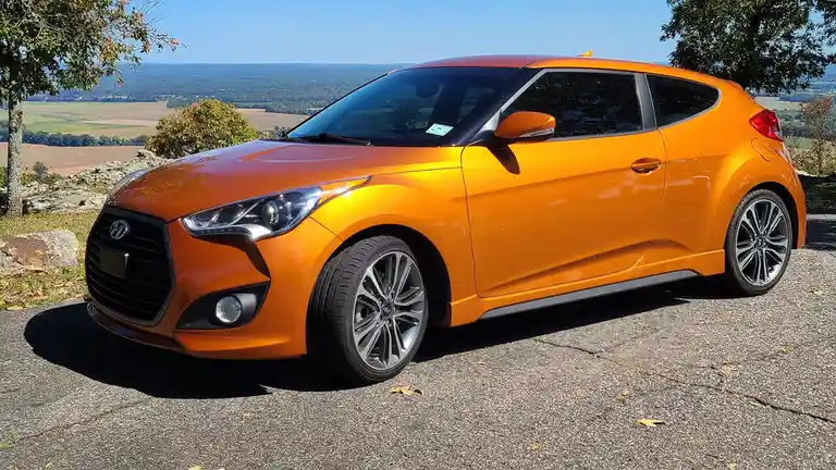 A bright orange Hyundai Veloster is parked on a paved overlook with a scenic view of rolling hills and trees under a clear…