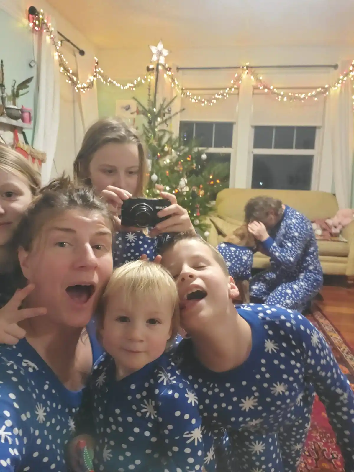A family in matching blue pajamas with white snowflake and polka dot patterns poses for a selfie in a living room decorated…