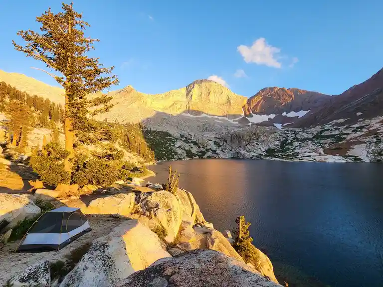 A serene mountain camping scene features a small gray and white tent pitched on rocky terrain beside a calm alpine lake. A…