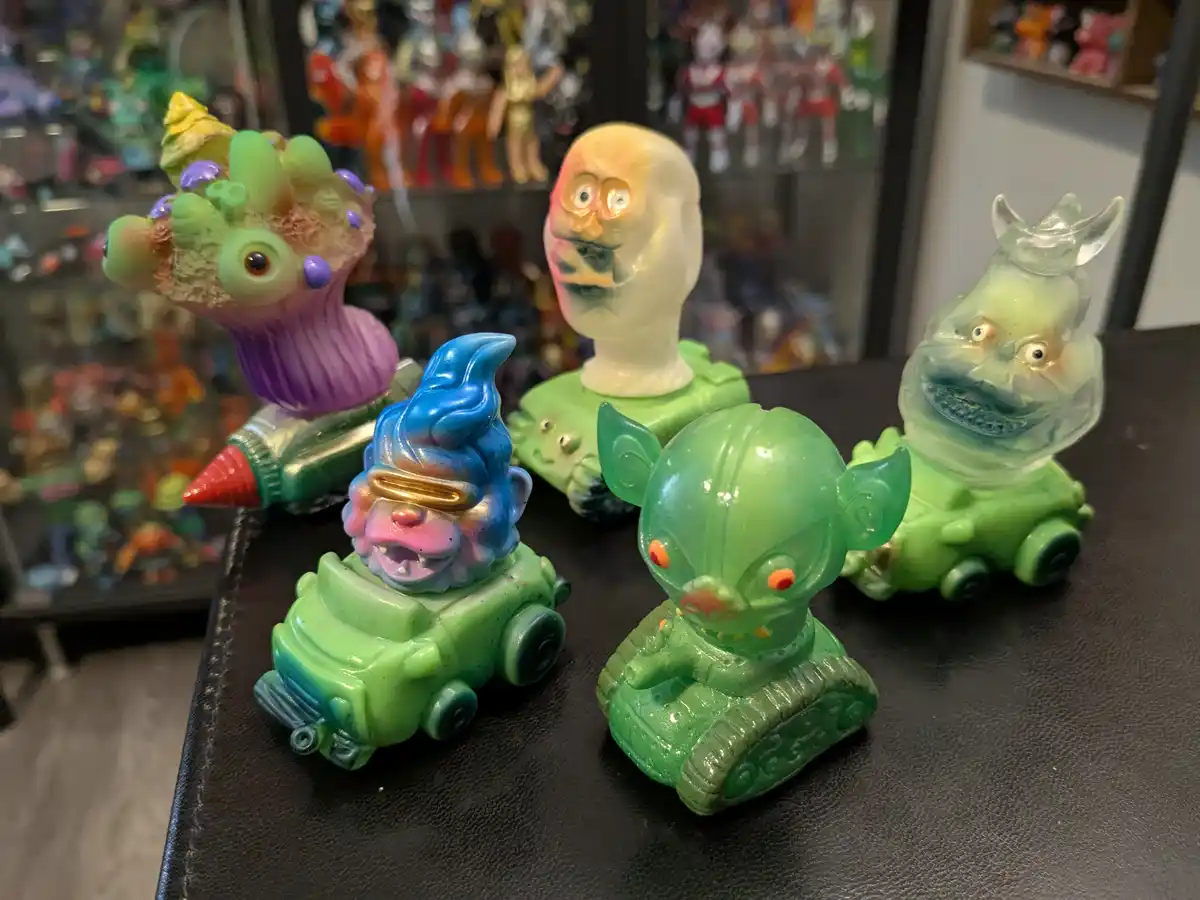 Five colorful, translucent, grotesque monster figurines with exaggerated facial features are arranged on a black surface…