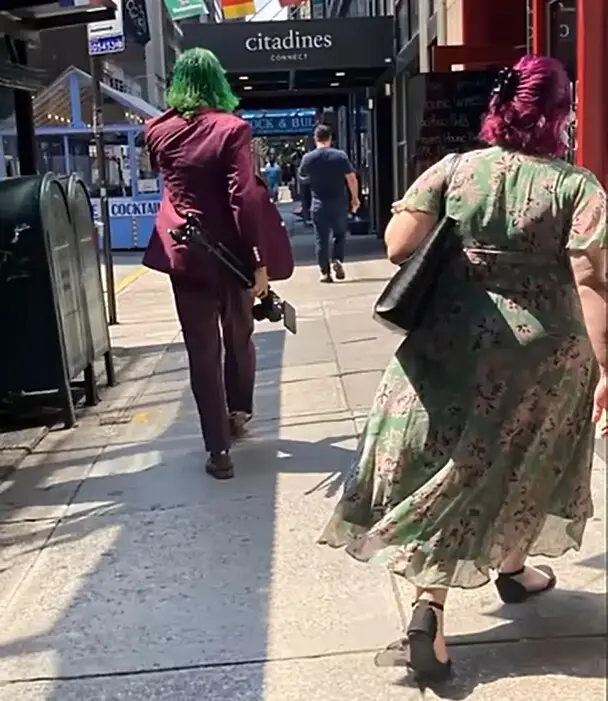 Two people walk away from the camera down a city sidewalk. The person on the left has bright green hair and wears a maroon…