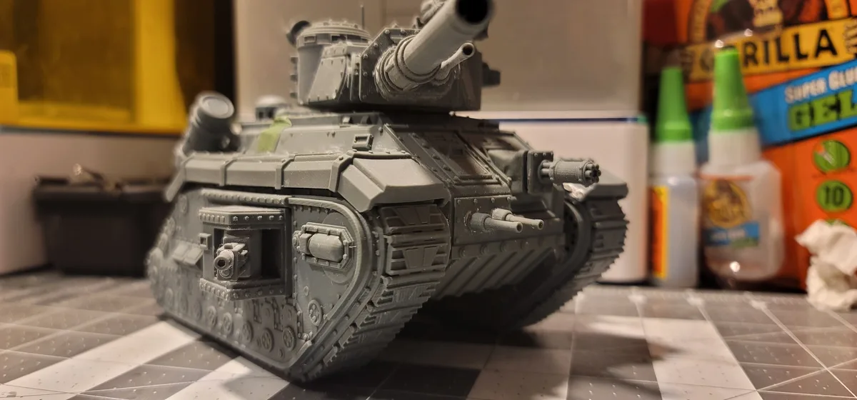 A gray, unpainted model tank sits on a checkered work mat, surrounded by hobby supplies like Gorilla Super Glue bottles. The…