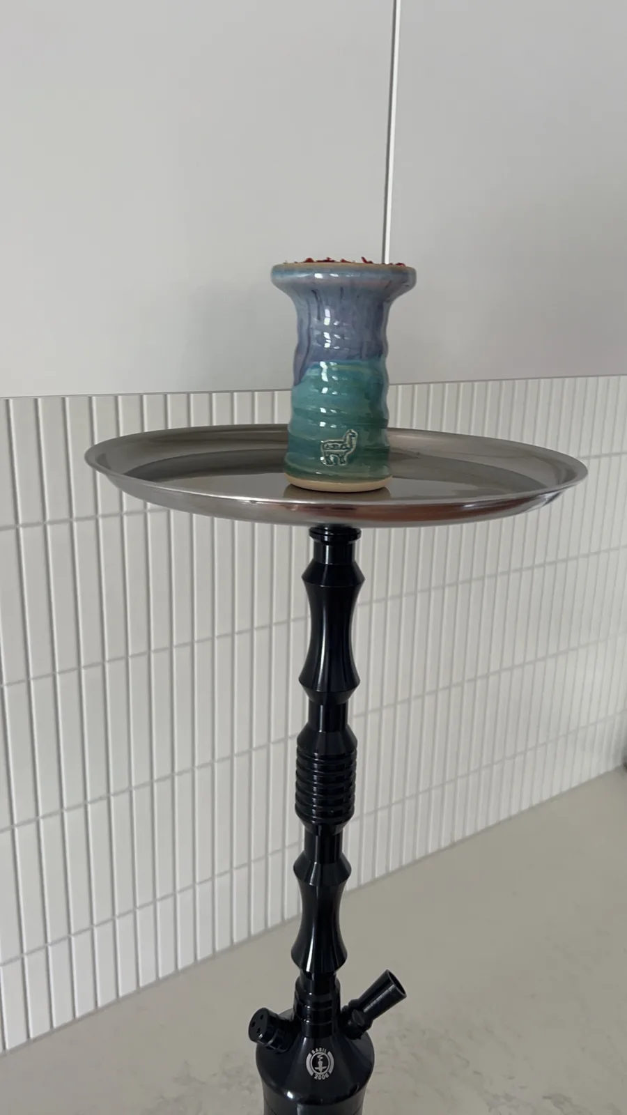 A hookah with a ceramic bowl shaped like a llama sits on its tray against a white tiled wall. The bowl is blue and green…