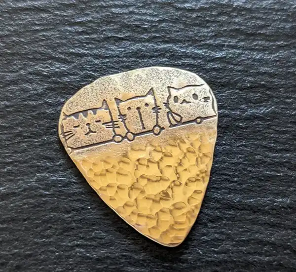 A golden, hammered metal guitar pick features three stylized cats engraved in a row, each sitting on a small wheel…