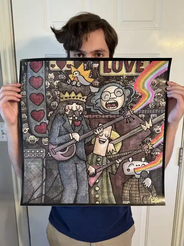 A person holds up a colorful, densely illustrated poster featuring cartoonish figures, including a crowned man with a…