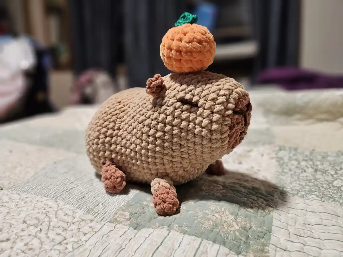 A handmade crocheted plush toy resembling a chubby, tan-colored rodent with a small orange pumpkin balanced on its head…