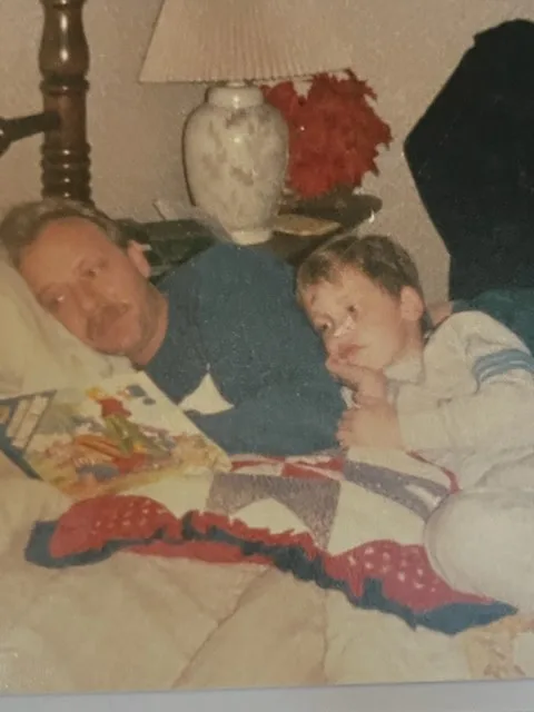 A grainy, old photograph shows a man and a young boy lying side-by-side on a bed under a patchwork quilt. The man, wearing a…