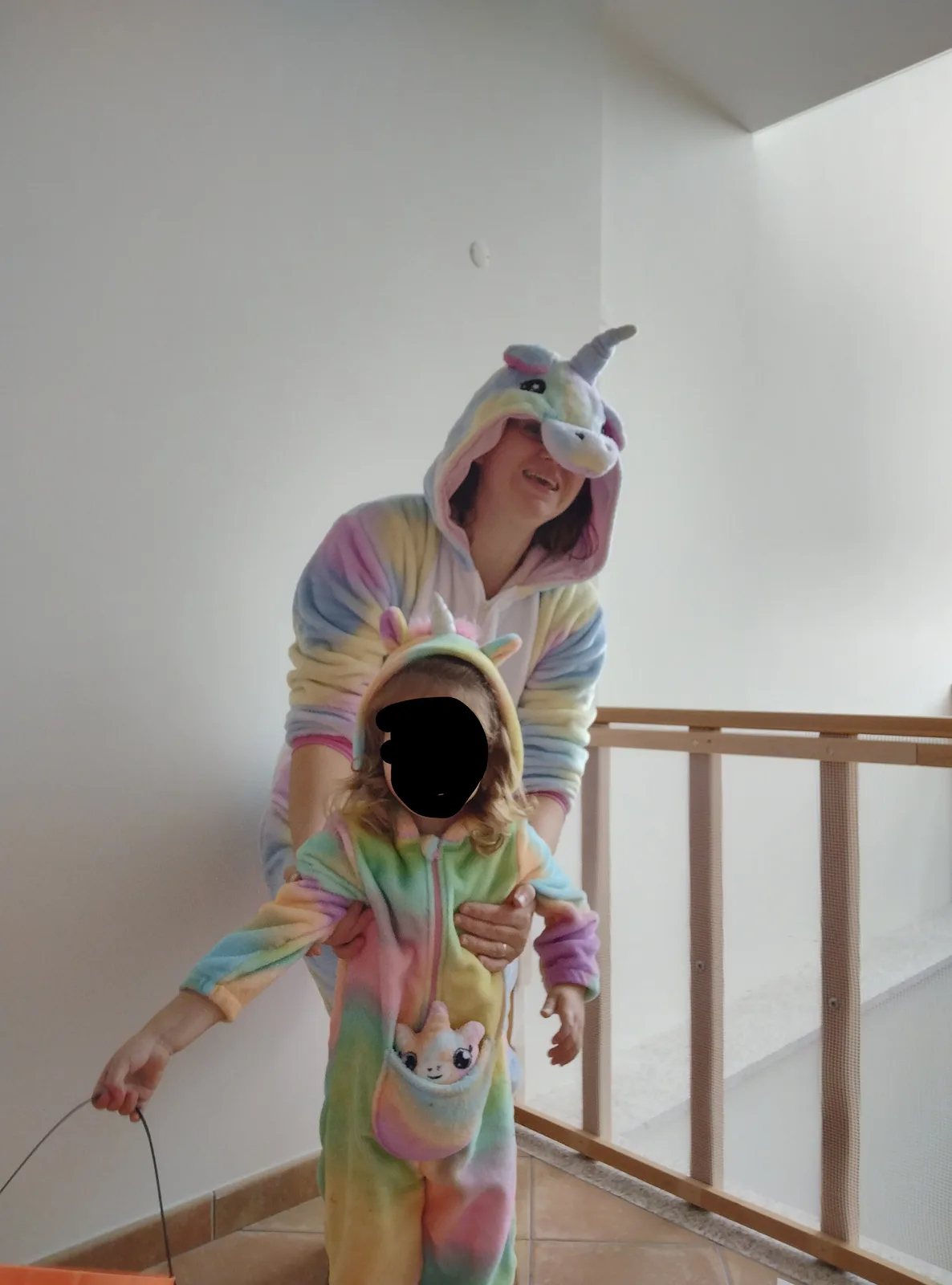 A woman and a small child, both wearing matching rainbow-colored unicorn onesies with horned hoods, stand on a staircase…