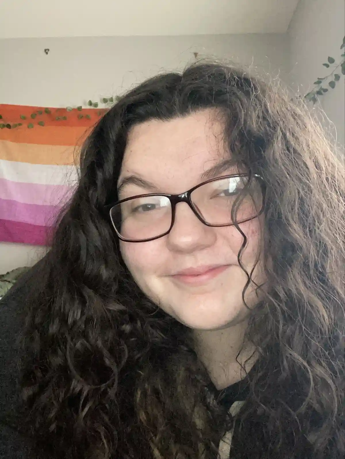 A person with long, dark curly hair and glasses smiles subtly while looking at the camera. Behind them, a rainbow-colored…