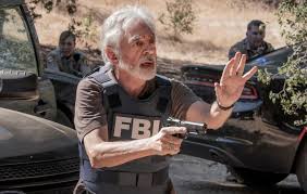 An older man with gray hair and a beard, wearing a tactical vest with 'FBI' printed on it, holds a handgun in one hand and…