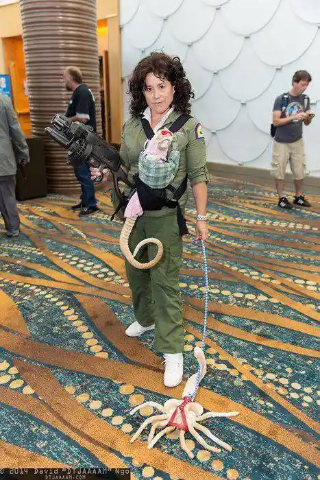 A woman dressed in a Ghostbusters uniform holds a proton pack and carries a baby in a carrier, while a plush ghost toy on a…
