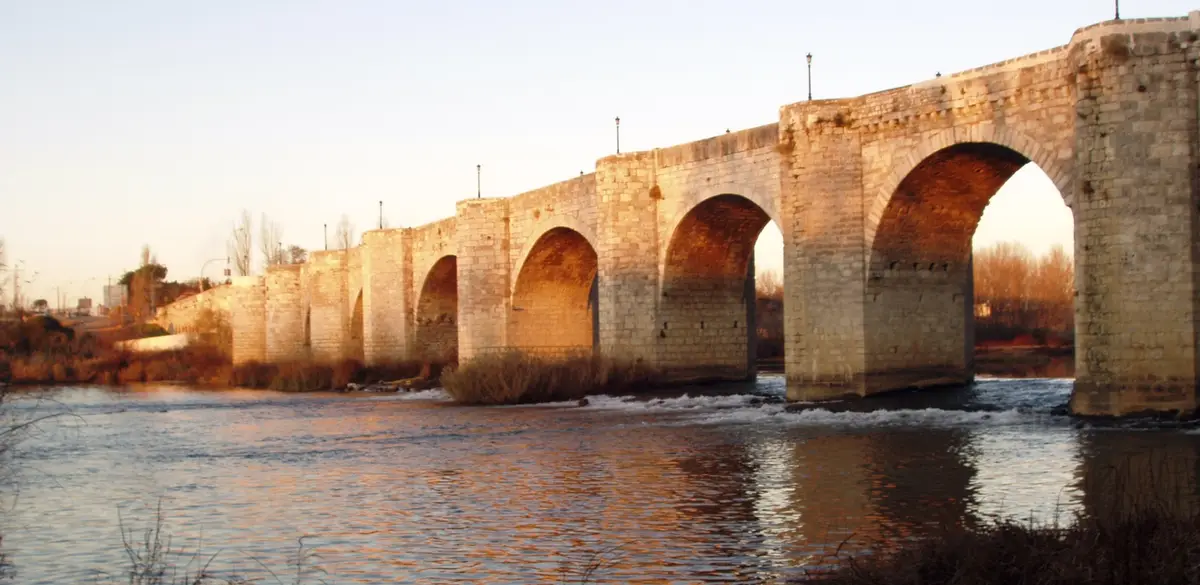 A scenic photo of a long, stone arch bridge spanning a river at sunset, with warm light reflecting on the water. The…