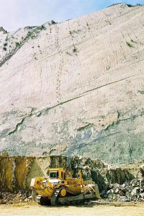 A yellow Caterpillar bulldozer sits at the base of a massive, steep, light-gray quarry wall. The wall shows horizontal…