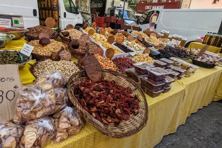 A vibrant outdoor market stall covered in a yellow tablecloth displays numerous baskets and bins of dried goods including…
