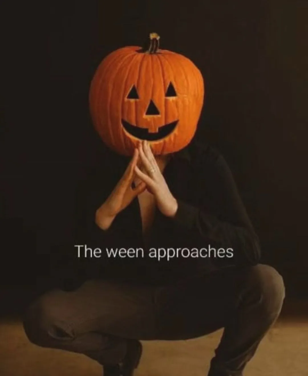A person crouching with hands clasped in prayer, wearing a carved jack-o'-lantern as a head covering against a dark…