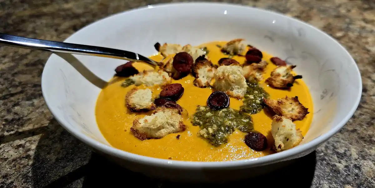 A white ceramic bowl filled with orange-colored soup, topped with toasted croutons, dark round garnishes, and green pesto…