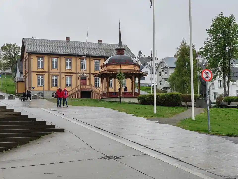 A wide shot of a public square with a yellow wooden building labeled 'Radsjua' and a gazebo, featuring pedestrians, a…