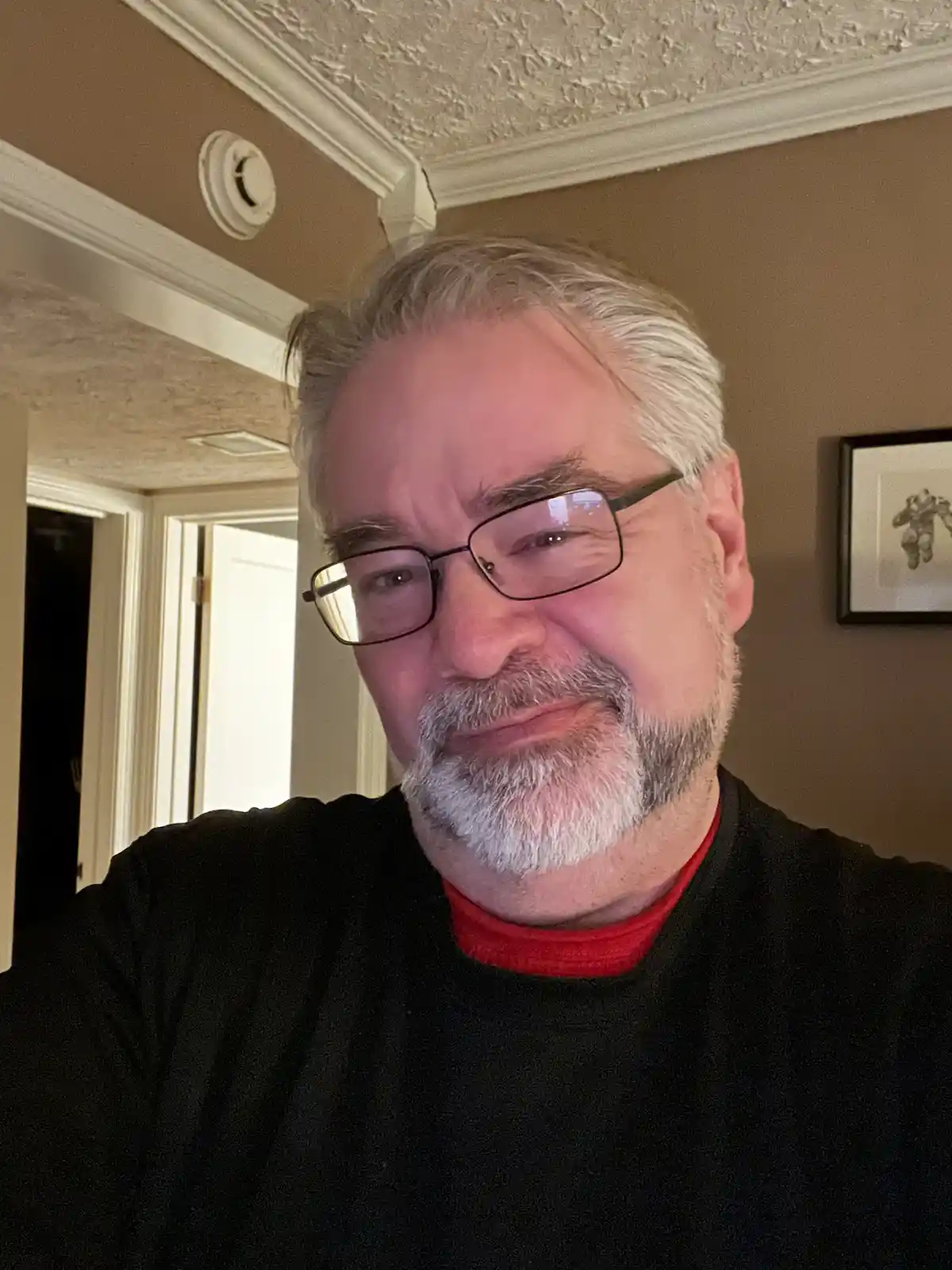 A middle-aged man with gray hair and glasses, wearing a black shirt over a red t-shirt, looks directly at the camera with a…