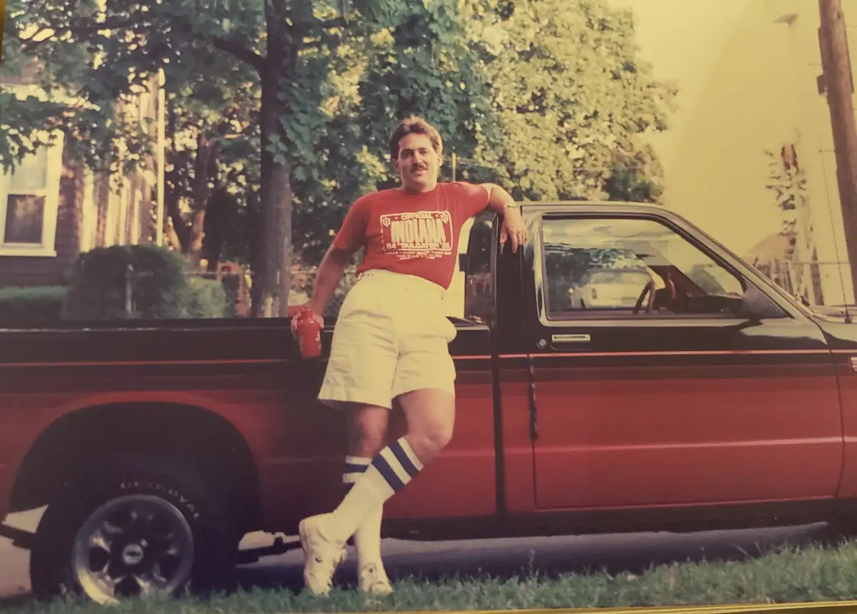 A man in a red 'Official Indiana 84 Tailgater 85' t-shirt, white shorts, and striped socks leans against a red pickup truck…