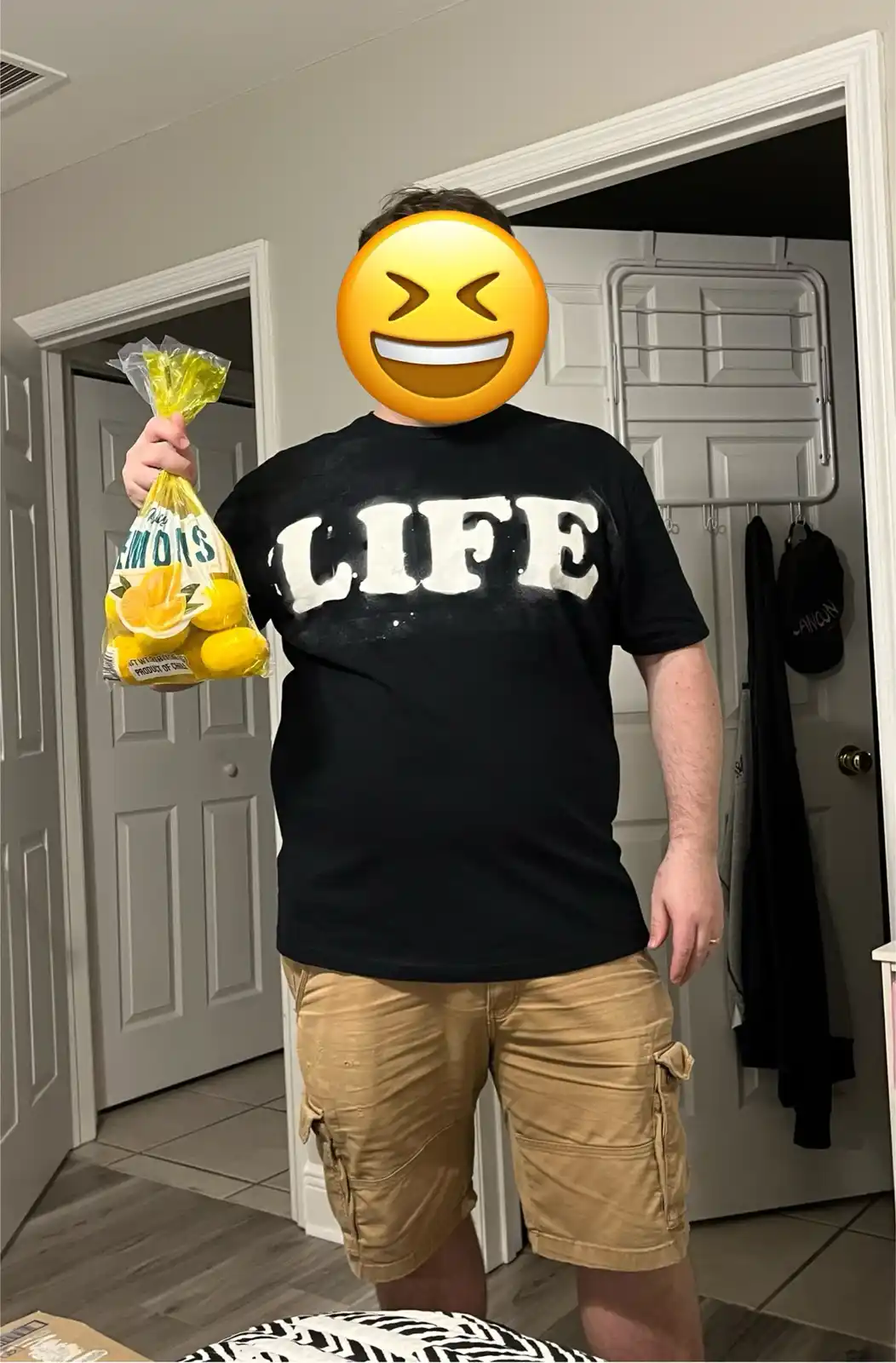 A person wearing a black t-shirt with the word 'LIFE' in large white letters stands indoors holding a plastic bag of lemons…