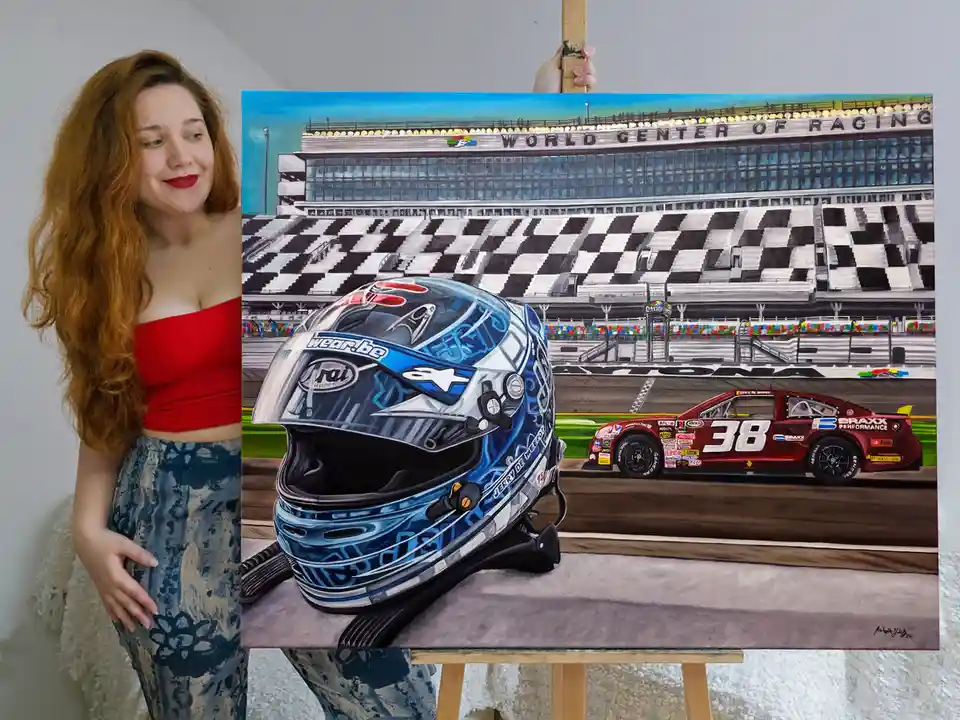 A woman with long red hair and red lipstick stands beside an easel displaying a painting of a blue and silver racing helmet…