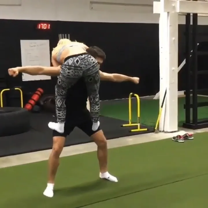 Two men are in a gym; one is standing with his arms outstretched while the other is on his back, also with arms…