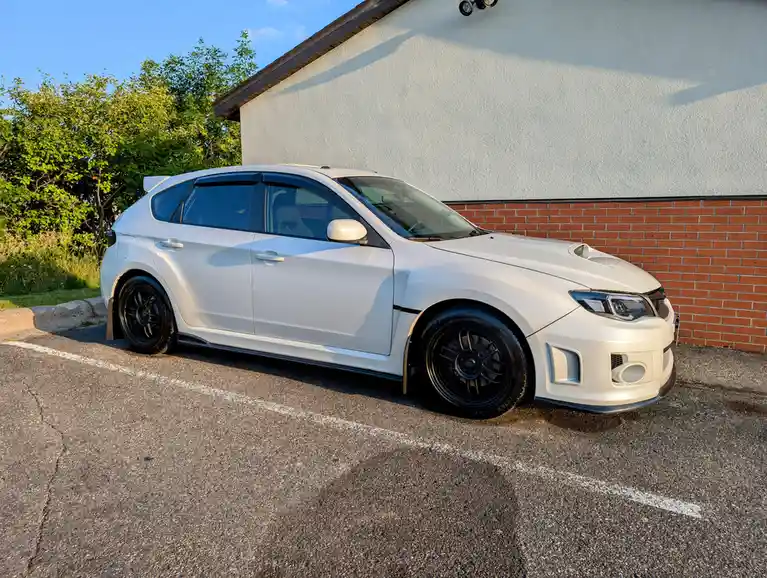 A white modified hatchback car with black wheels and aftermarket body kits is parked in a lot next to a brick wall. The…