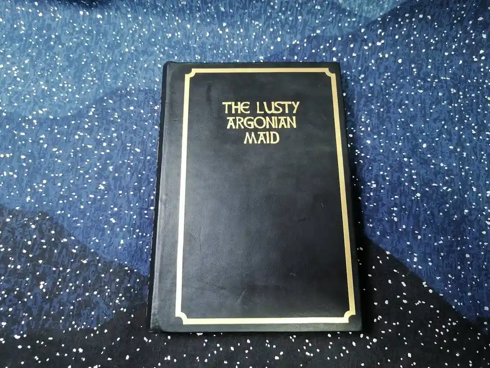 A black book with gold lettering and border sits on a blue and black speckled fabric. The title on the cover reads 'THE…