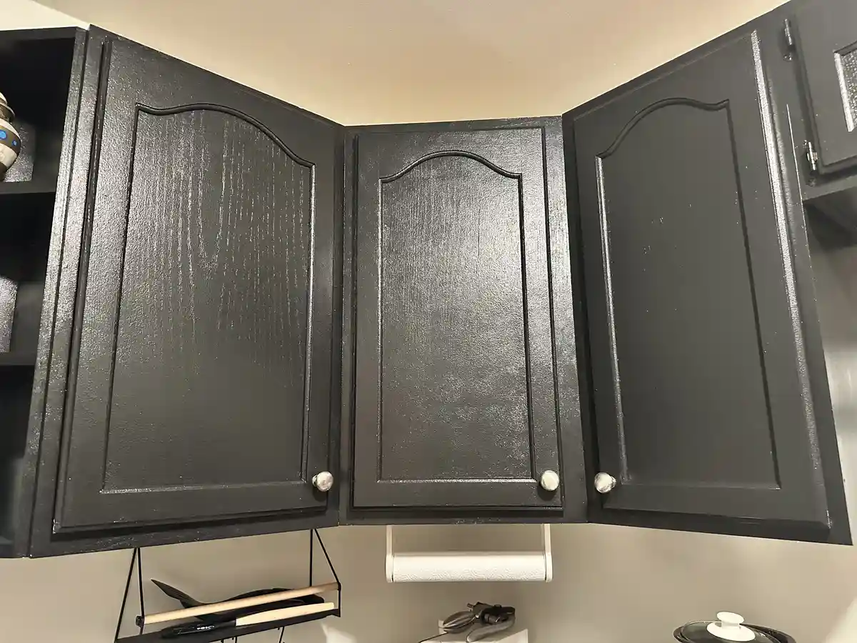 A photo shows three black-painted kitchen cabinets with silver knobs, all slightly ajar, revealing a beige wall behind…