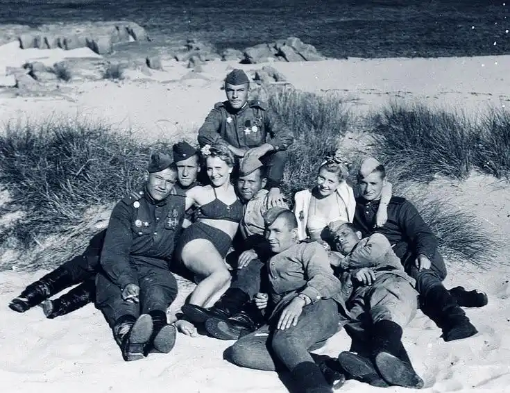 a vintage black-and-white photo shows a group of soldiers in uniform posing on a sandy beach with two women in swimsuits…