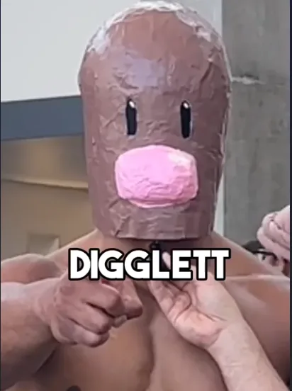 A shirtless person is wearing a handmade papier-mâché mask resembling a brown, round creature with two black slit eyes and a…