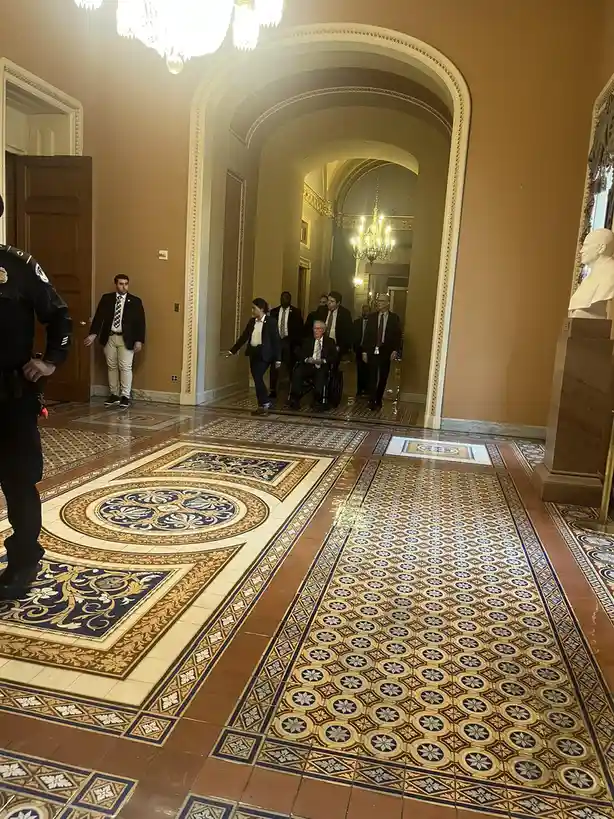A group of people, including a man in a wheelchair, are being escorted through an ornate hallway with patterned tile floors…