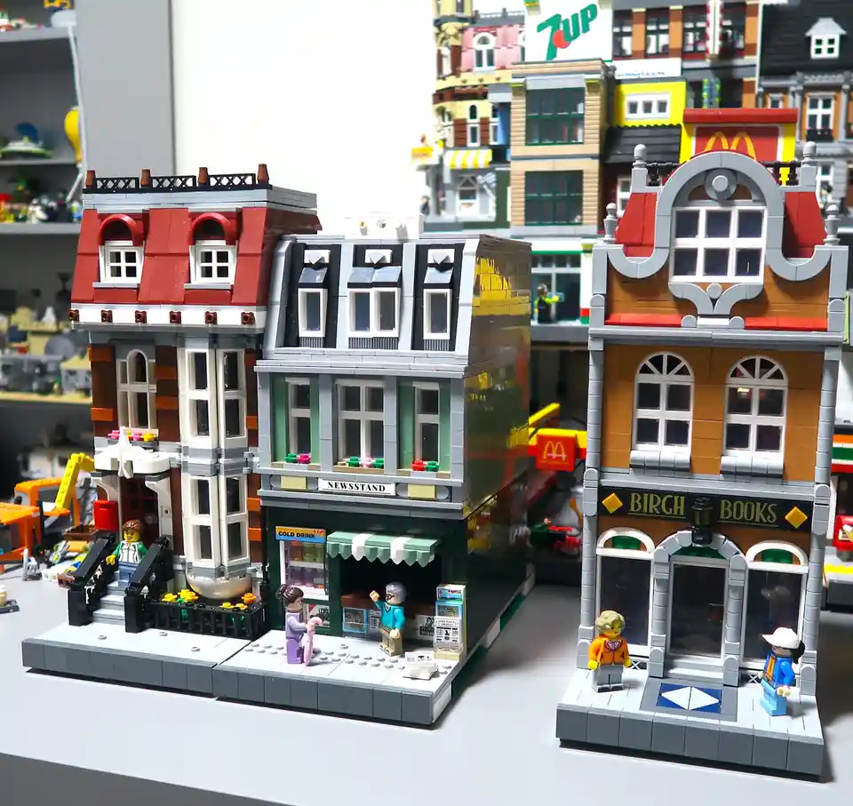A detailed LEGO cityscape featuring three connected buildings with minifigures, including a newsstand, a bookstore named…