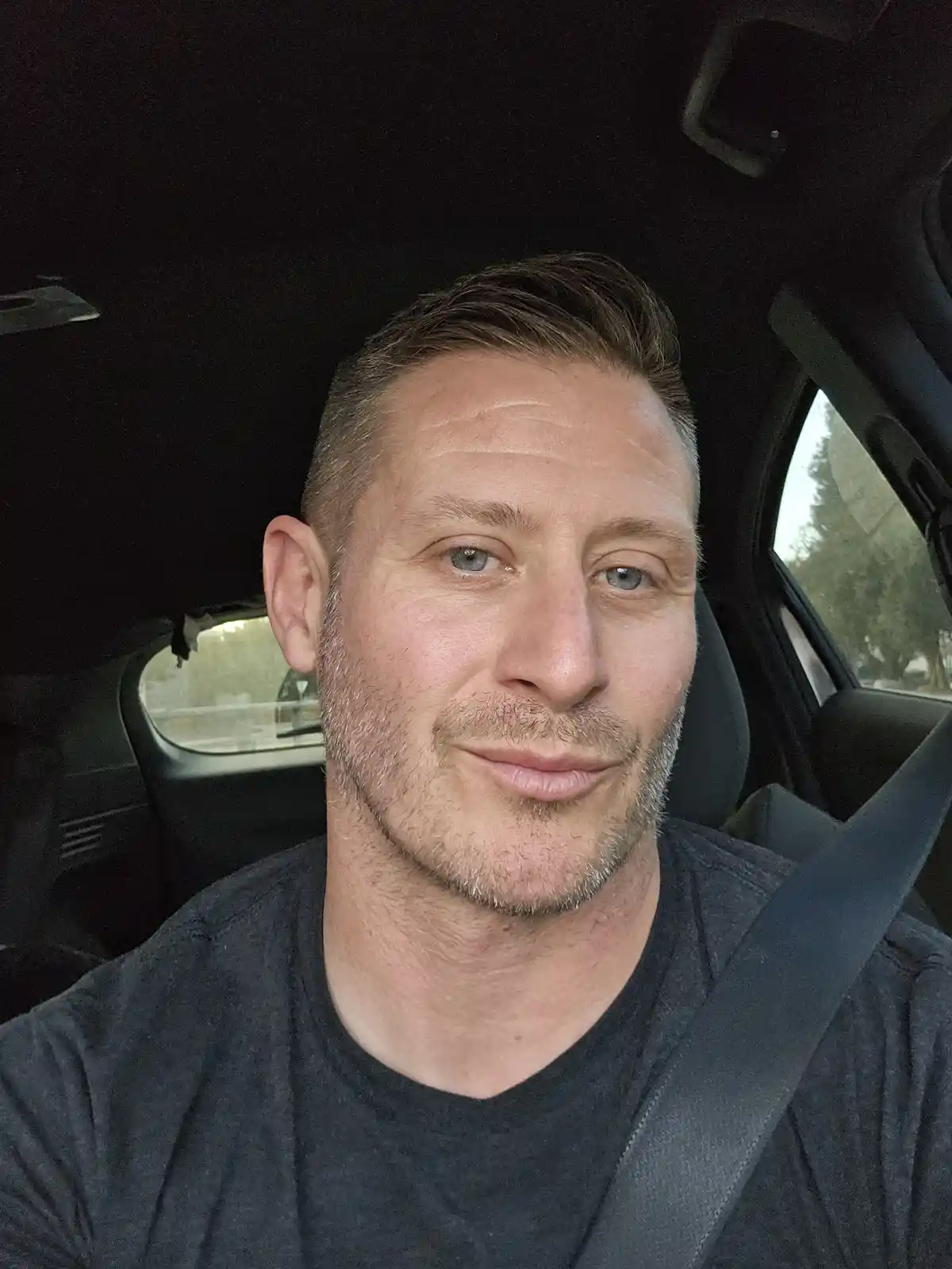 A man with short hair and a light beard sits in the driver's seat of a car, wearing a dark t-shirt and a seatbelt, looking…