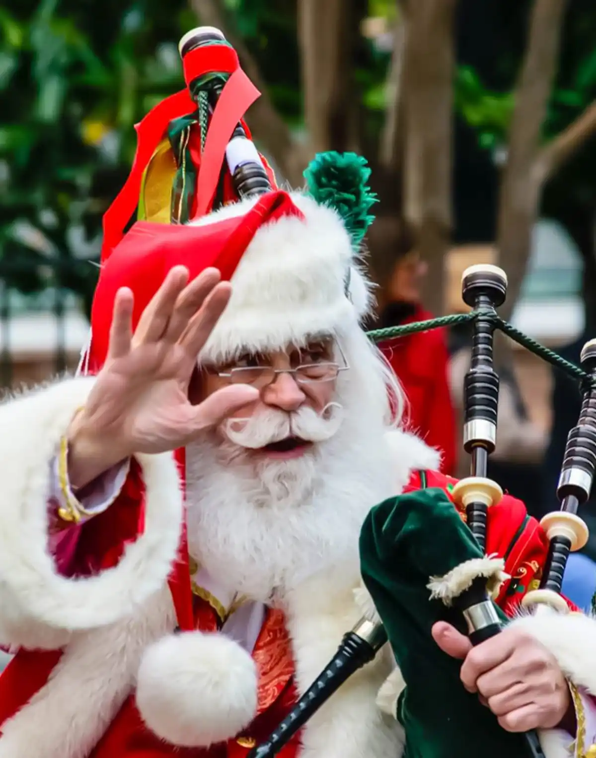 A man dressed as Santa Claus, wearing a red and white fur-trimmed suit and hat, is playing a set of bagpipes while waving…