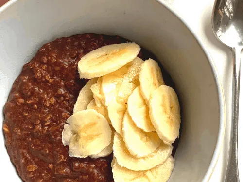 A close-up photo of a white bowl filled with dark brown oatmeal topped with a pile of sliced bananas, with a metal spoon…