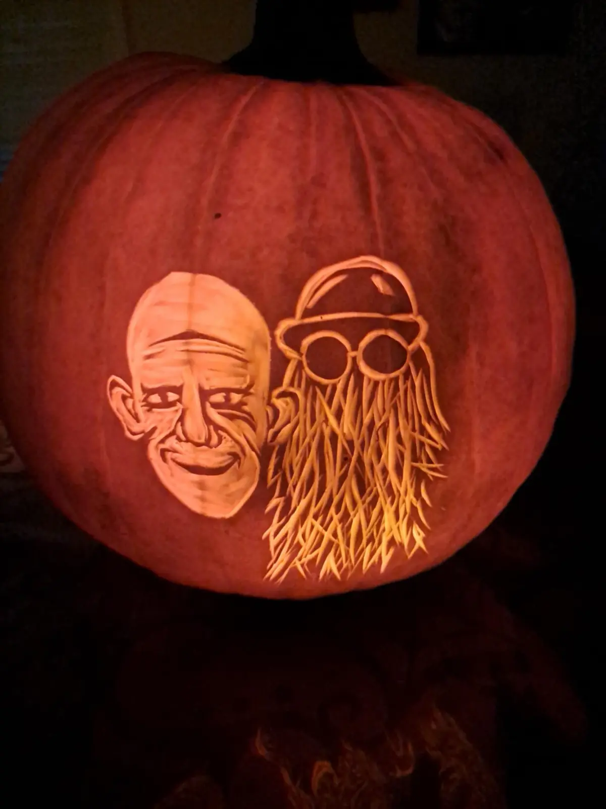 A carved pumpkin illuminated from within, featuring two distinct faces: one bald with a smiling expression and deep…