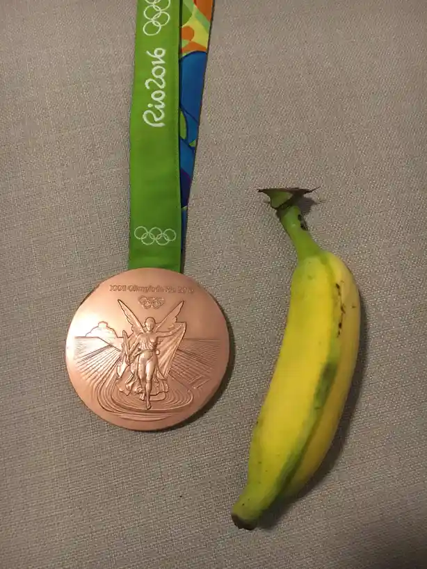 a rio 2016 olympic bronze medal with its green and colorful ribbon lies next to a yellow banana on a gray fabric surface…