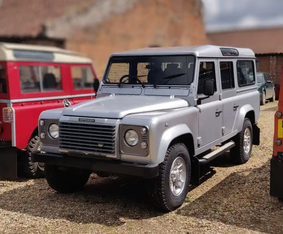 A silver Land Rover Defender is parked on gravel in front of a red Land Rover, with brick buildings in the background. The…