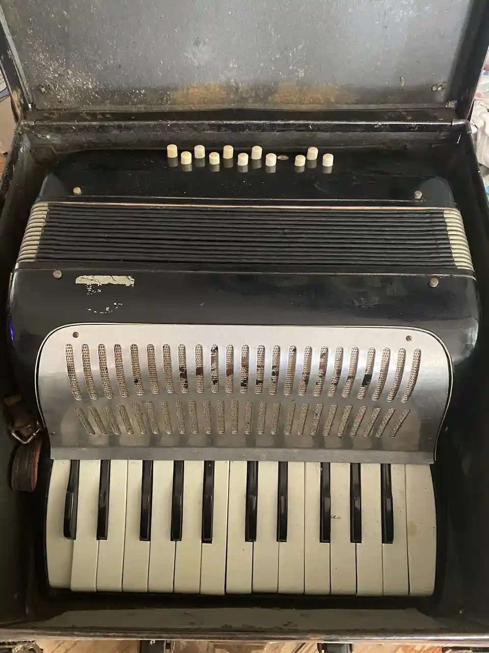 A close-up photo of a vintage, worn black accordion with a silver grille and piano-style keys, showing visible signs of age…