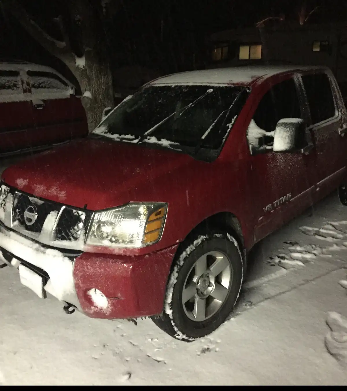 A red Nissan Titan pickup truck is parked outdoors at night, completely covered in snow on its roof, windows, mirrors, and…
