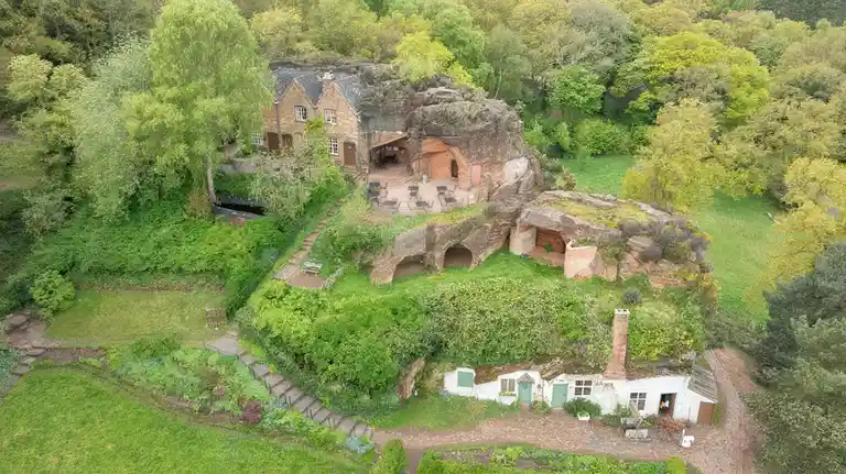 An aerial view shows a lush, green hillside with several buildings, including stone cottages and cave-like structures carved…