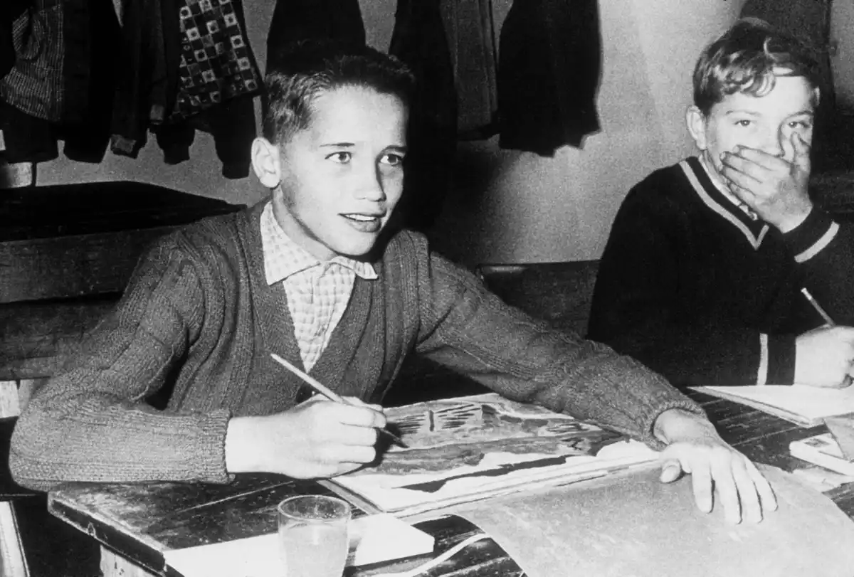 A black-and-white photo shows two young boys at a wooden desk. The boy on the left, wearing a cardigan over a checkered…