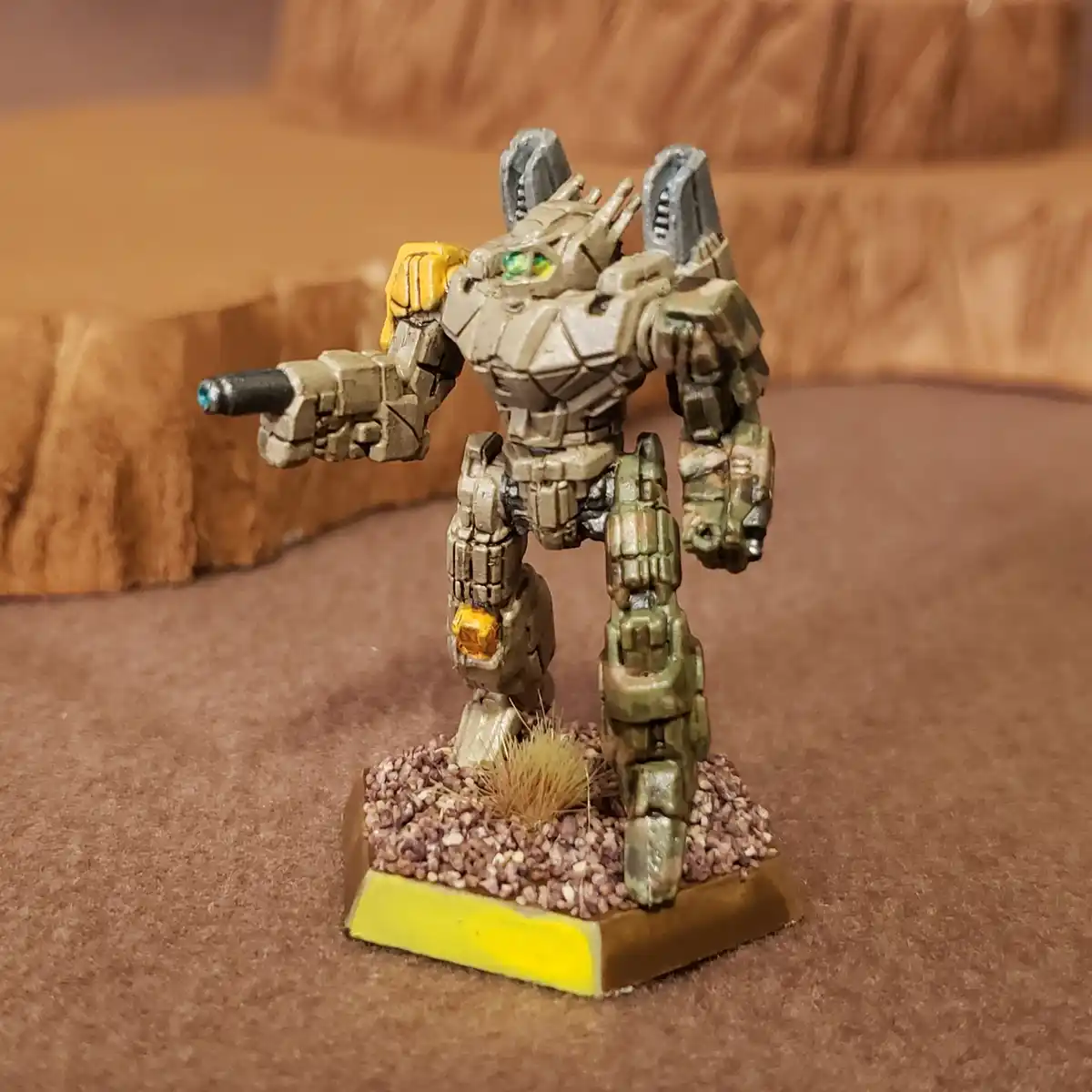 A painted miniature robot figurine stands on a base with gravel and a small tuft of grass, holding a gun and looking…