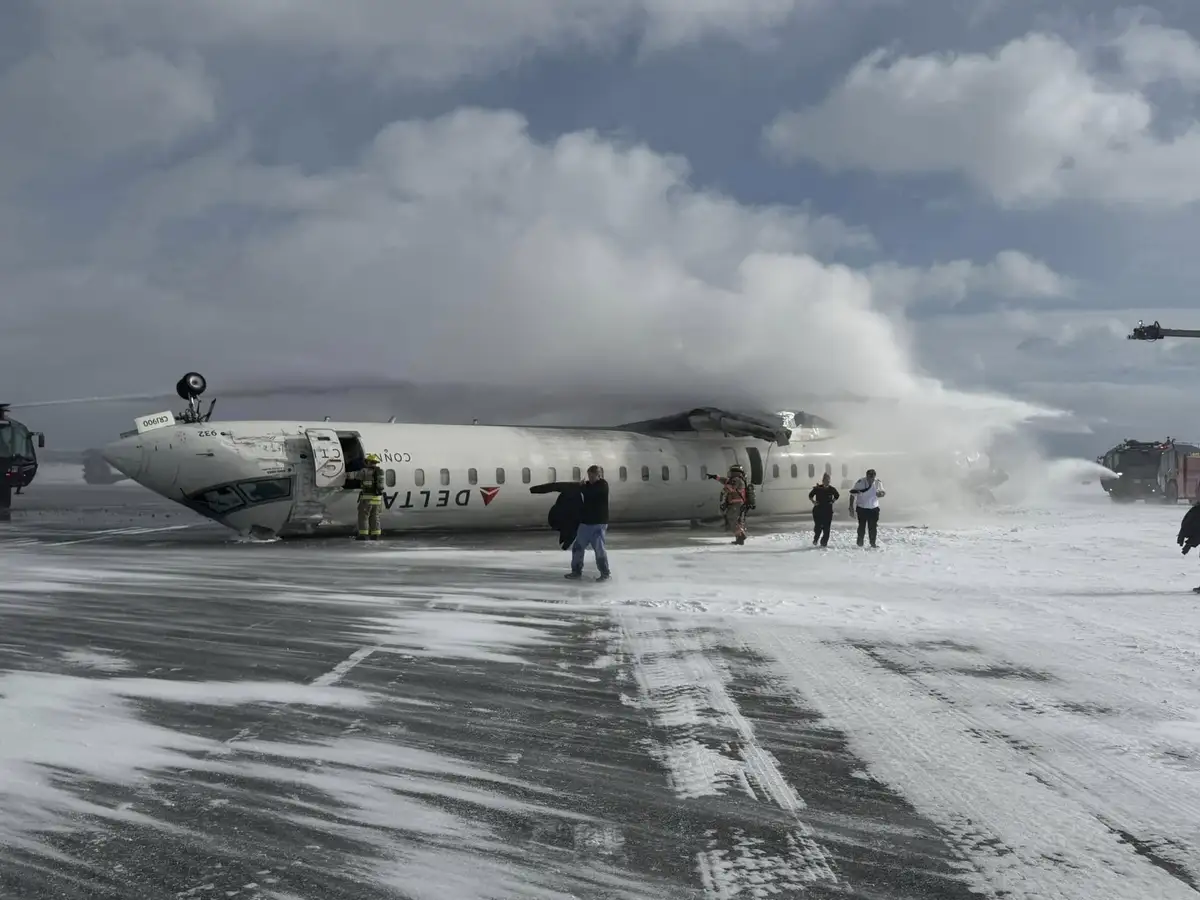 A Delta airplane is on a snowy runway, visibly damaged and being sprayed with water by fire trucks. Several emergency…