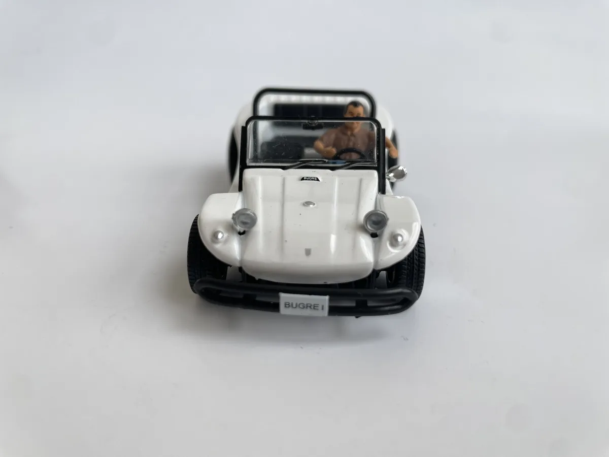 A white toy car with black trim and a small figurine driver is photographed from the front on a plain white surface. The…